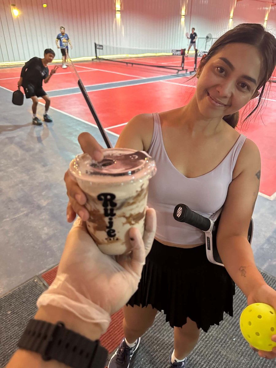 A Ruje Café drink being handed to a pickleball player beside the court, a game going on behind her
