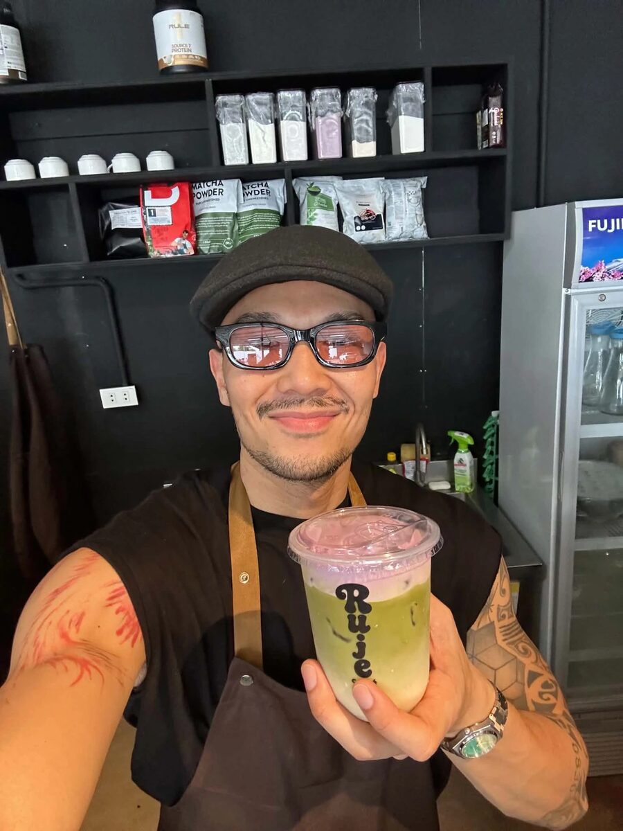 Russ, co-owner of Ruje Café, holding up an iced matcha with a purple foam top behind the counter